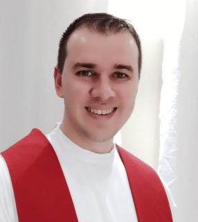 A imagem ilustra um padre católico, vestindo túnica branca e estola vermelha.