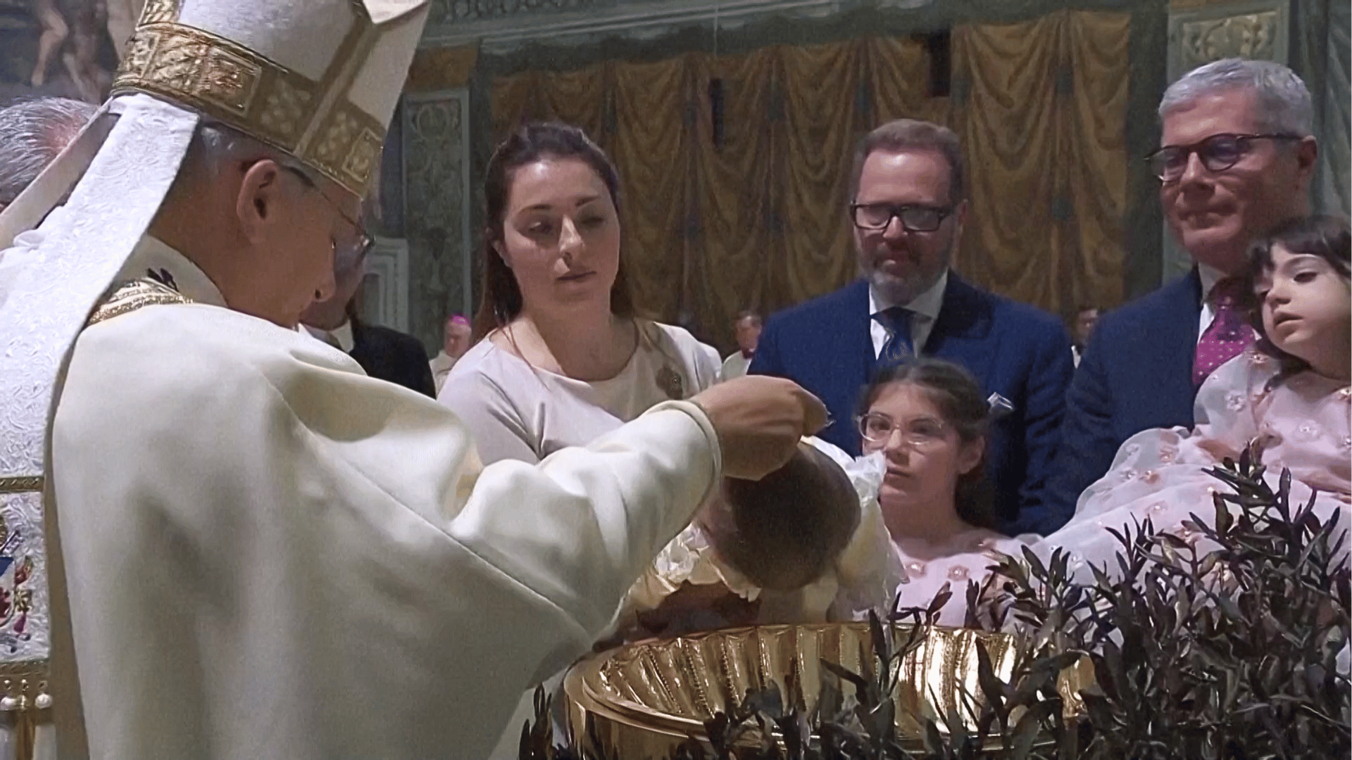 A imagem ilustra o Papa Leão XIV, liturgicamente paramentado, batizando um bebê. Membros da família da criança estão ao redor, observando o momento.
