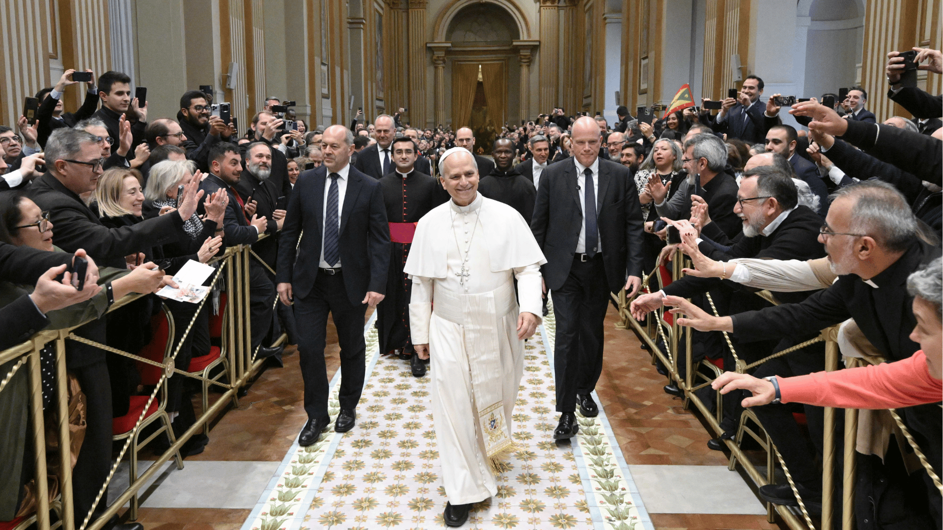 Papa caminha pelo corredor de uma sala do Vaticano, sorrindo e cumprimentando fiéis que se aproximam e registram o momento com celulares