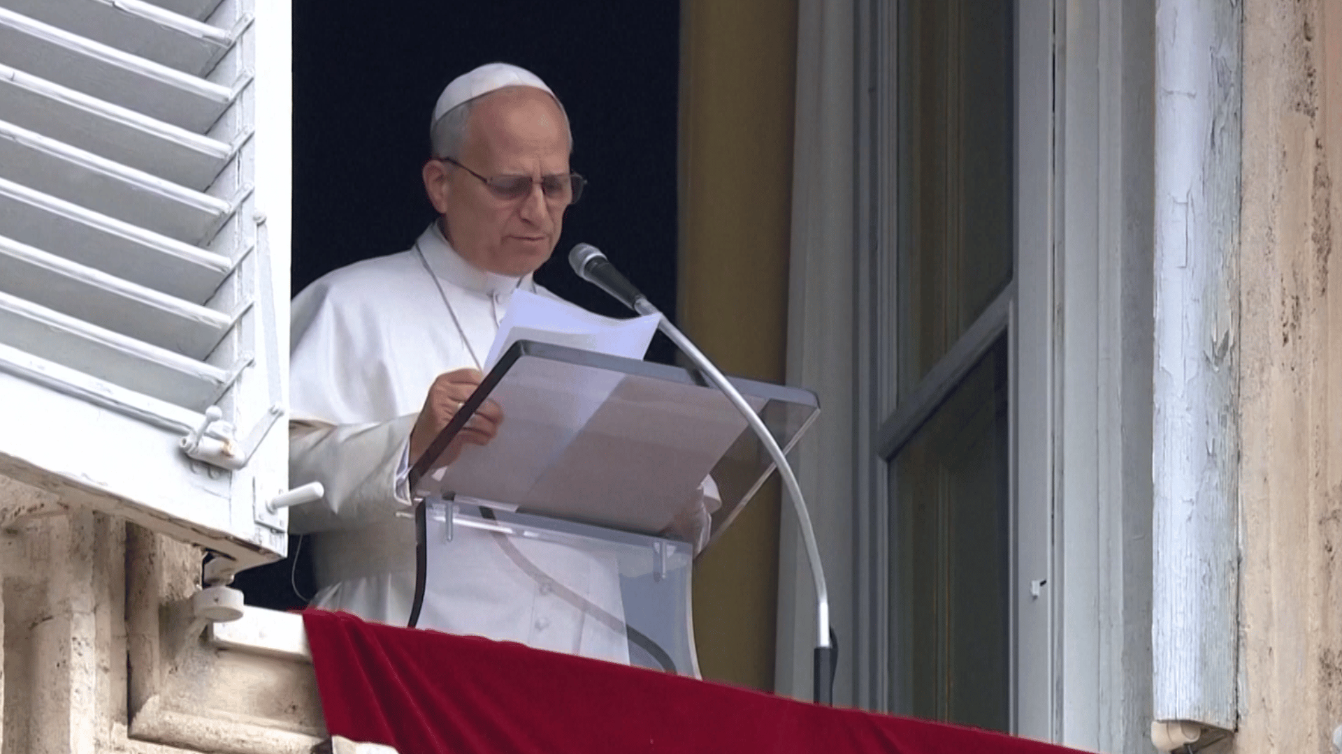A imagem ilustra o Papa Leão XIV durante a oração do Angelus. Ele econtra-se diante do parapeito de uma das janelas do Palácio Apostólico, no Vaticano, com suas habtiausi vestes pontifícias. O Santo Padre lê algumas folhas de papel que segura sobre um púlpito, diante de um microfone.