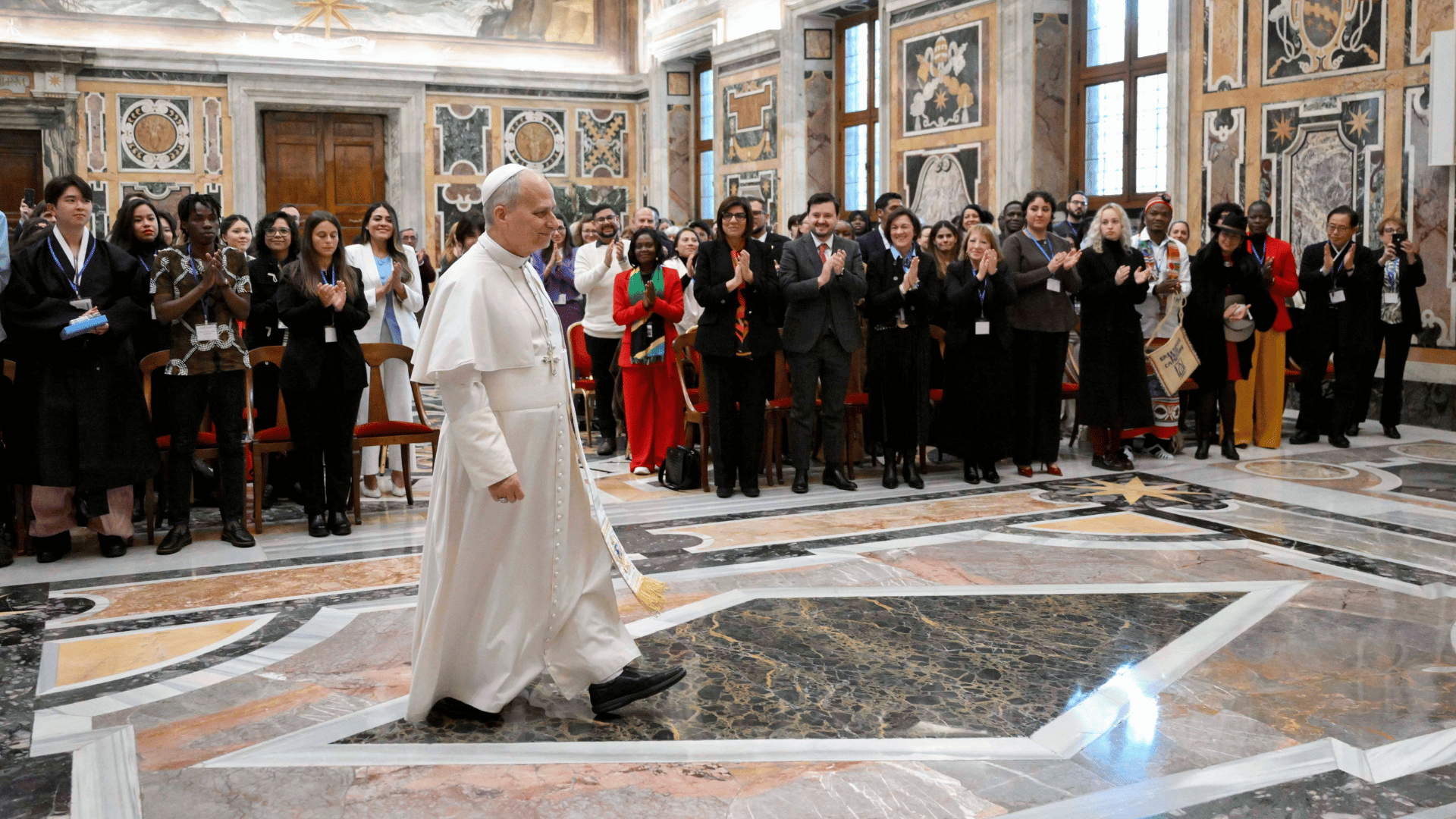 Papa Leão XIV é acolhido por participantes da audiência A imagem ilustra o Papa Leão XIV, com suas habituais vestes potifícias, caminhando enquanto adentra em uma sala. Ao fundo, vê-se diverssas pessoas aplaudindo-o em gesto de acolhida.