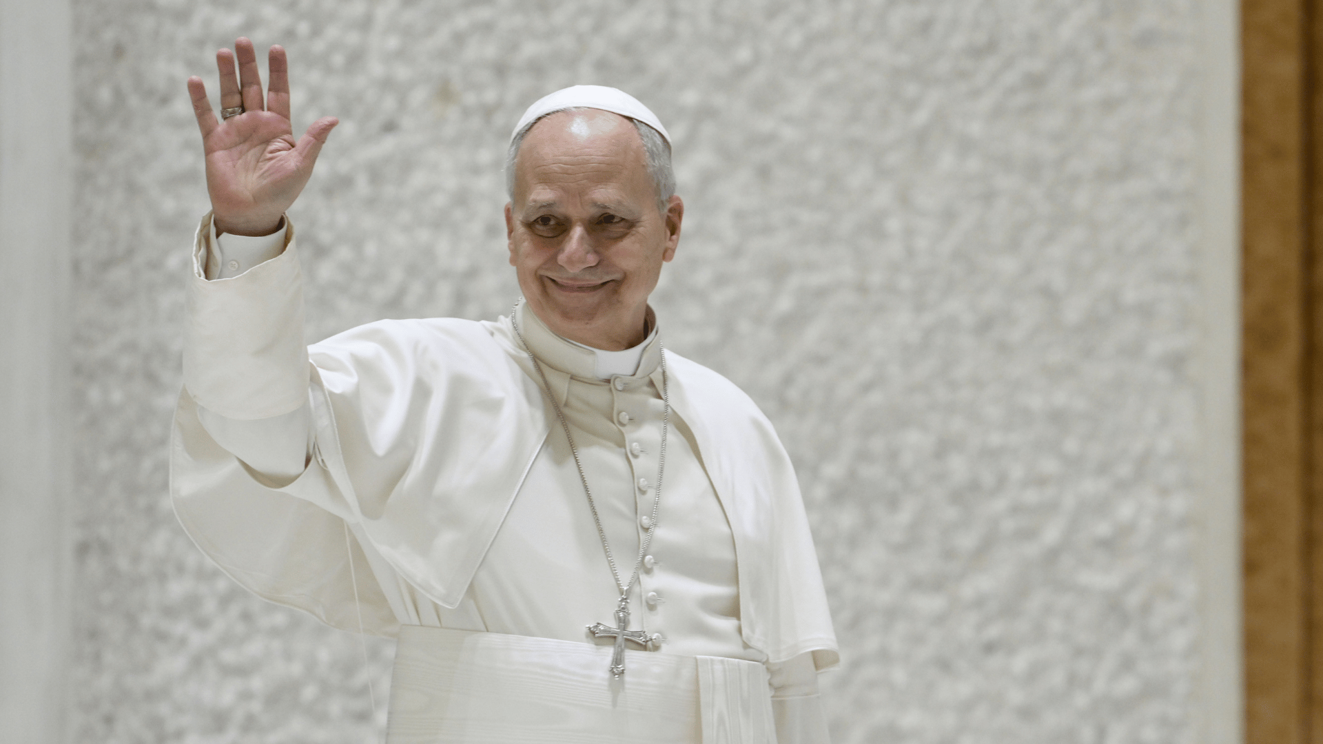 A imagem ilustra o Papa Leão XIV com suas habituais vestes pontifícias e o braço direito erguido em saudação.