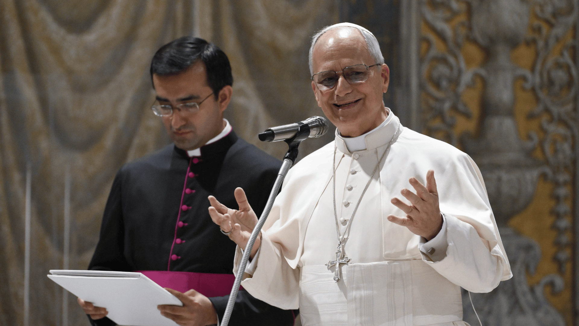 A imagem ilustra o Papa Leão XIV em suas habituais vestes pontifícias. Ele está em primeiro plano na foto, com as mãos erguidos à altura do peito, gesticulando enquanto fala ao microfone. Ao seu lado direito, em segundo plano, está um monsenhor que tem pele branca e cabelos pretos e lisos, olhando para uma pasta que segura em suas mãos.