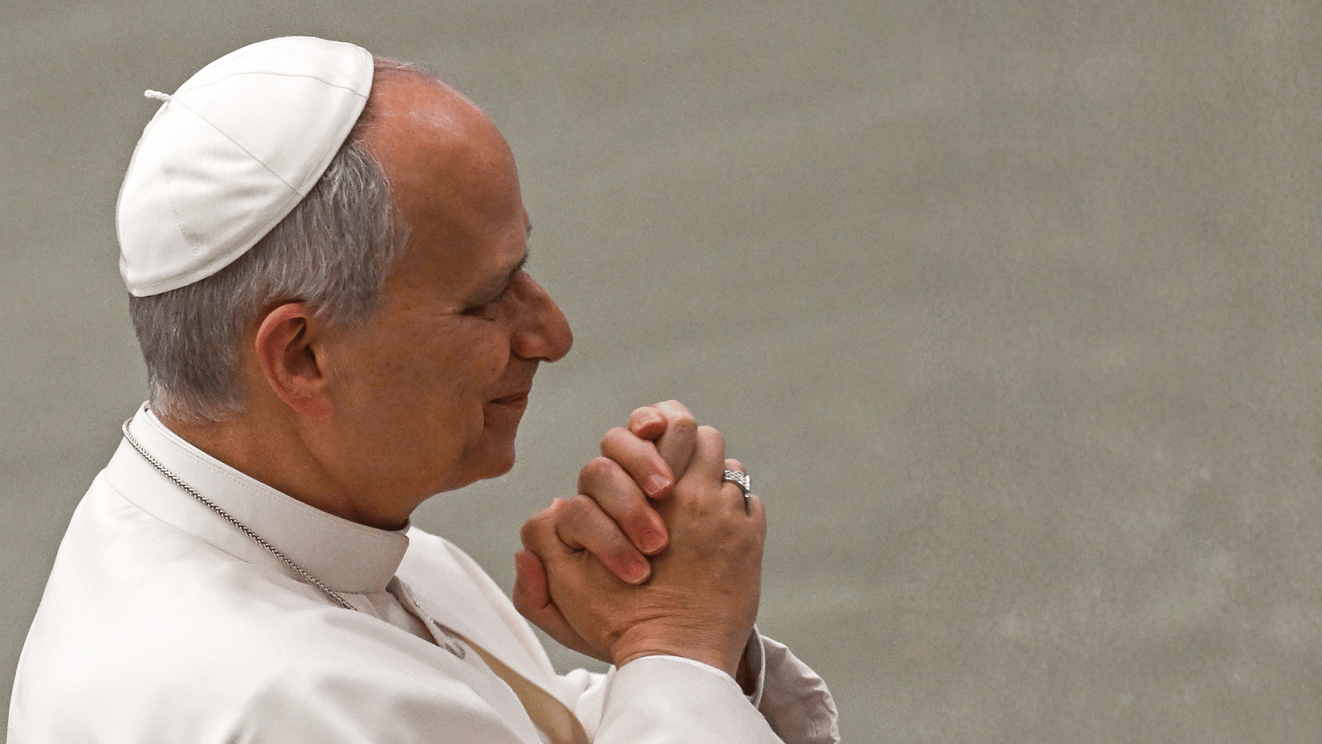 A imagem ilustra o Papa Leão XIV com suas habituais vestes pontifícias e mãos juntas, de perfil.