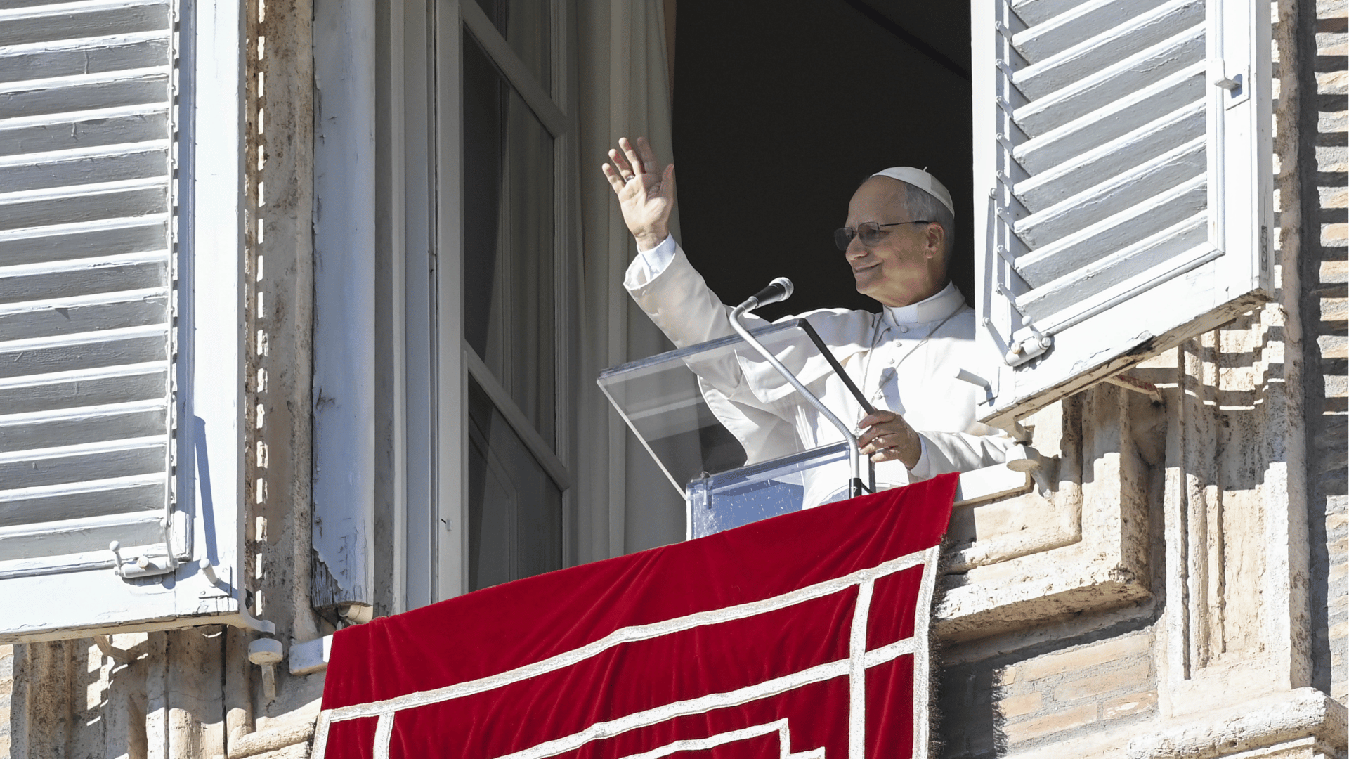 Papa vestido de branco acena para os fiéis a partir da janela do Palácio Apostólico do Vaticano, falando ao microfone atrás de um púlpito, com a bandeira do Vaticano pendurada na sacada.