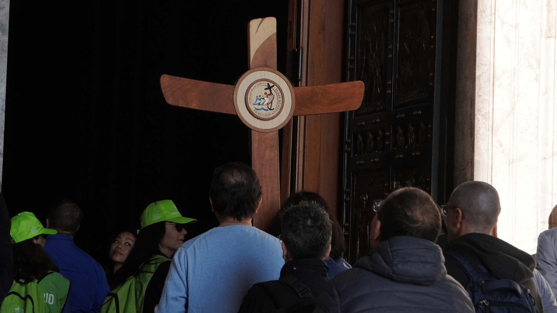 Peregrinos seguram a cruz jubilar e passam pela porta santa da basílica de são pedro