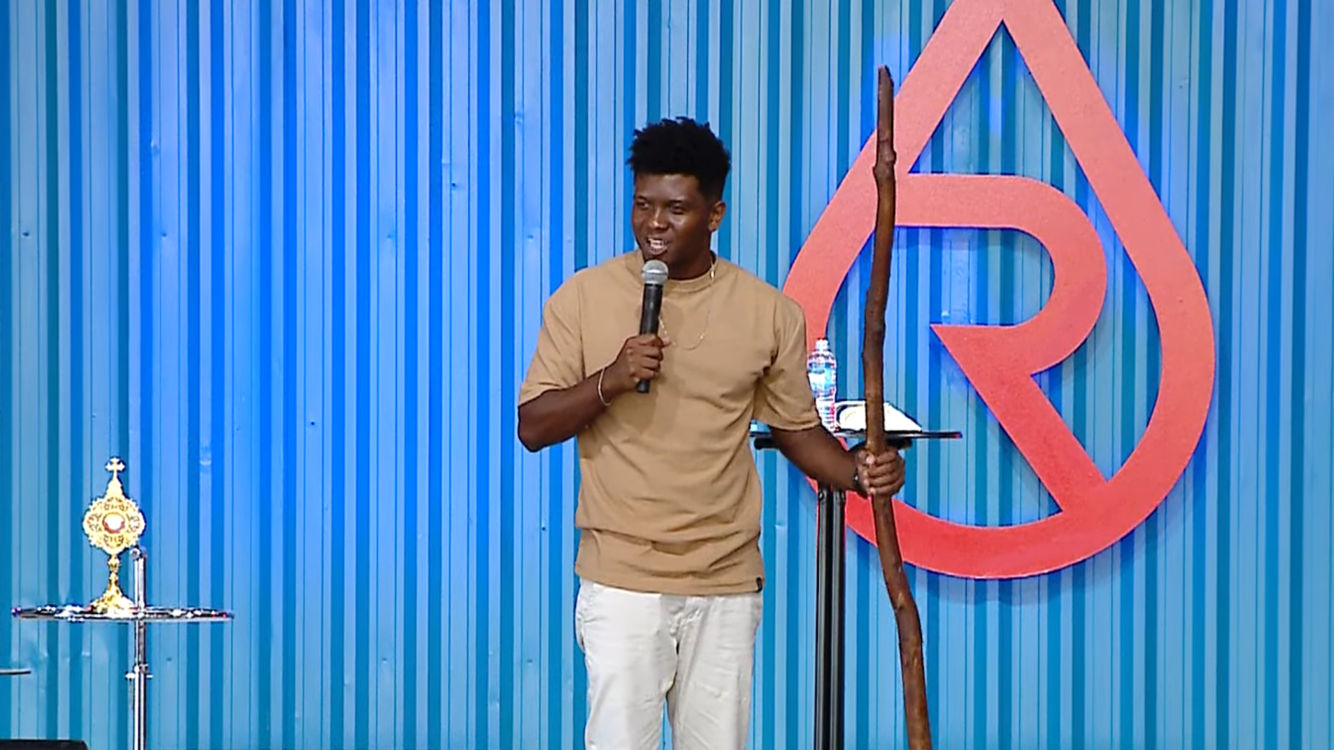 Rick no Acampamento Revolução Jesus 2026 A imagem ilustra um home de pele negra e cabelo preto, vestido com uma camisa bege e cala branca. Ele segura um microfone com a mão esquerda e um cajado de madeira com a mão direita. Ao funo, vê-se uma parede azul com o símbolo do Revolução Jesus, vermelho, afixado, e, à esquerda, um relicário dourado.