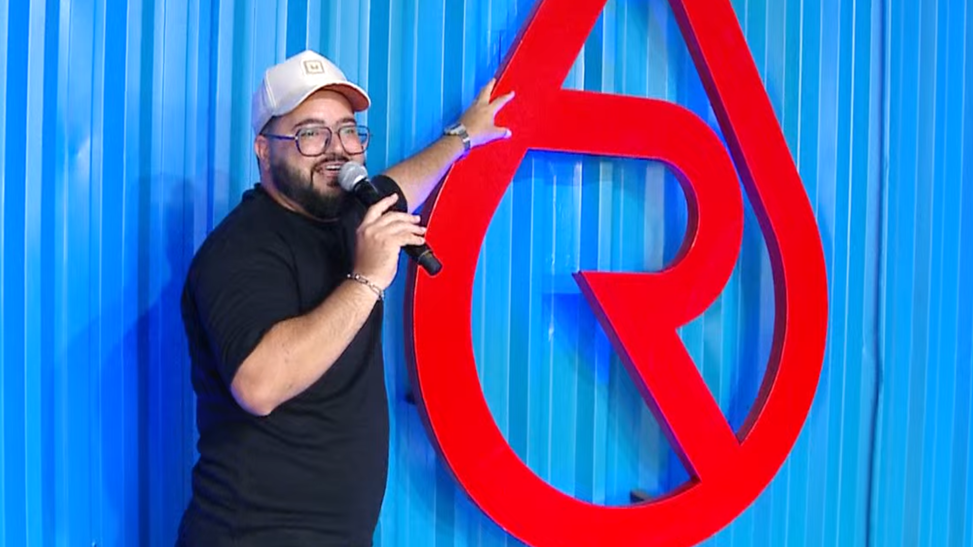 Rodrigo Carpanelli no Acampamento Revolução Jesus 2026 A imagem ilustra um homem de pele branca e bigode e barba pretos, vestindo uma camisa preta e boné branco. Ele fala diante de um microfone, em sua mão direita, enquanto com a esquerda aponta o símbolo do Revolução Jesus, vermelho, afixado em uma parede azul.