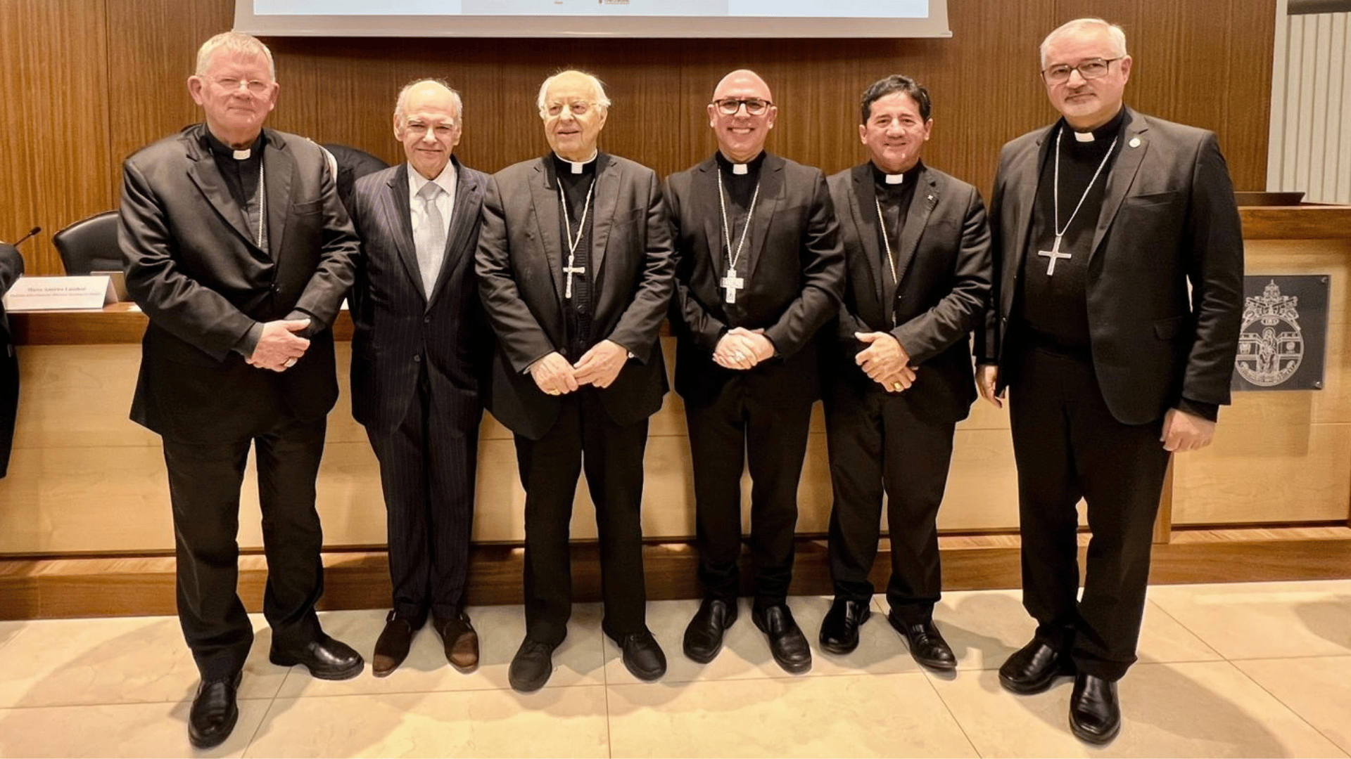 A imagem ilustra uma fila de seis homens conferencistas, dos quais cinco vestem clérgima e paletó preto e um veste um paletó comum.