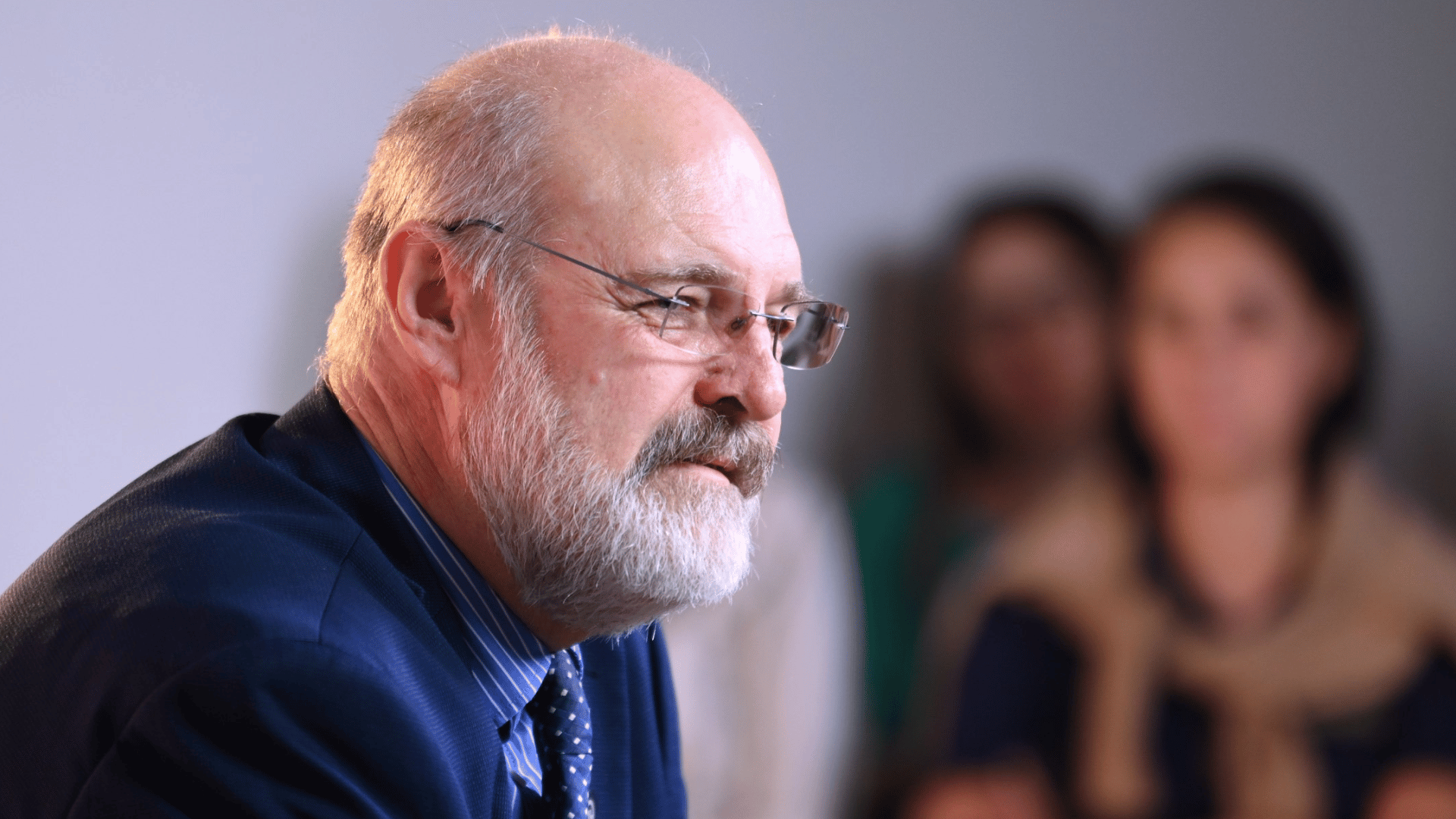 Silvonei José A imagem ilustra um homem de pele branca, com cabelos grisalhos ralos e barba grisalha, de perfil. Ao fundo vê-se algumas pessoas observando-o.