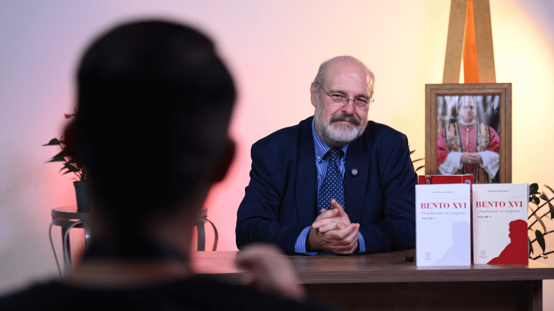 Silvonei José ouve a pergunta de um participante A imagem ilustra um homem de pele branca, com cabelos grisalhos ralos e barba grisalha, vestido com um paletó azul marinho. Ele escuta um jovem de cabelos pretos que está à sua frente, em primeiro plano.