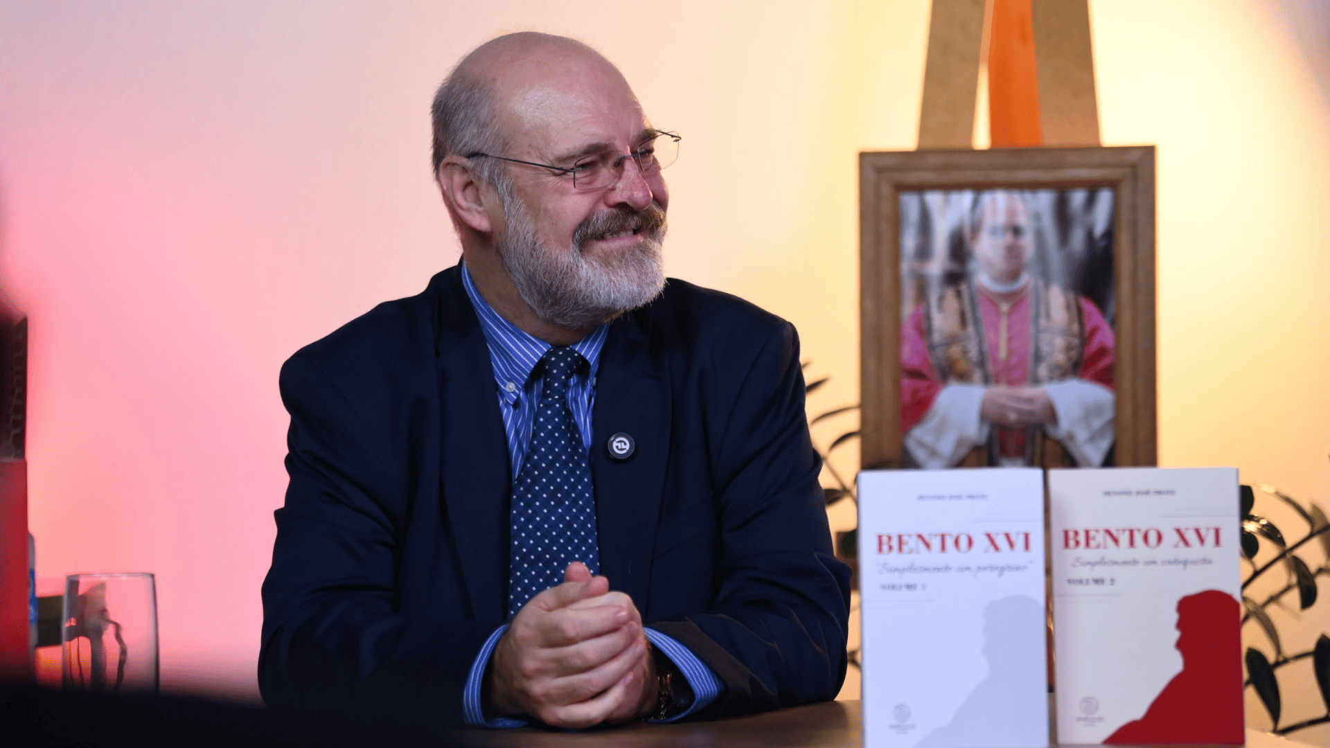 Silvonei José escreveu dois livros sobre o Papa Bento XVI e prepara o terceiro A imagem ilustra um homem de pele branca, com cabelos grisalhos ralos e barba grisalha, vestido com um paletó azul marinho. Ele está sentado, com os braços debruçados em uma mesa à sua frente. Nela estão dois livros com a inscrição "Bento XVI" e capa branca. Ao fundo vê-se um quadro com uma foto do Papa Leão XIV.