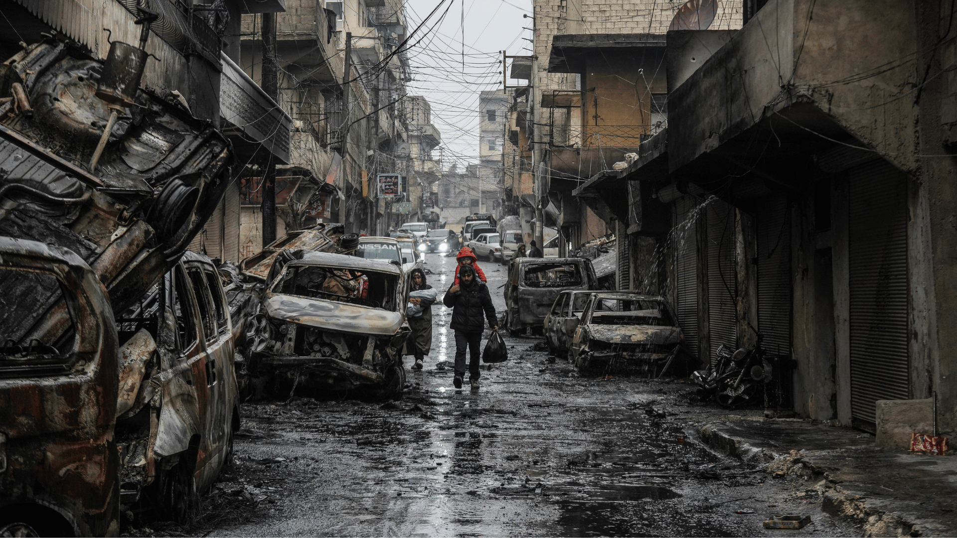 Moradores de Aleppo caminham por rua destruída por conflito A imagem ilustra uma rua com diversos carros queimados e prédios atingidos por explosões. Ao centro, uma família caminha em meio à destruição.