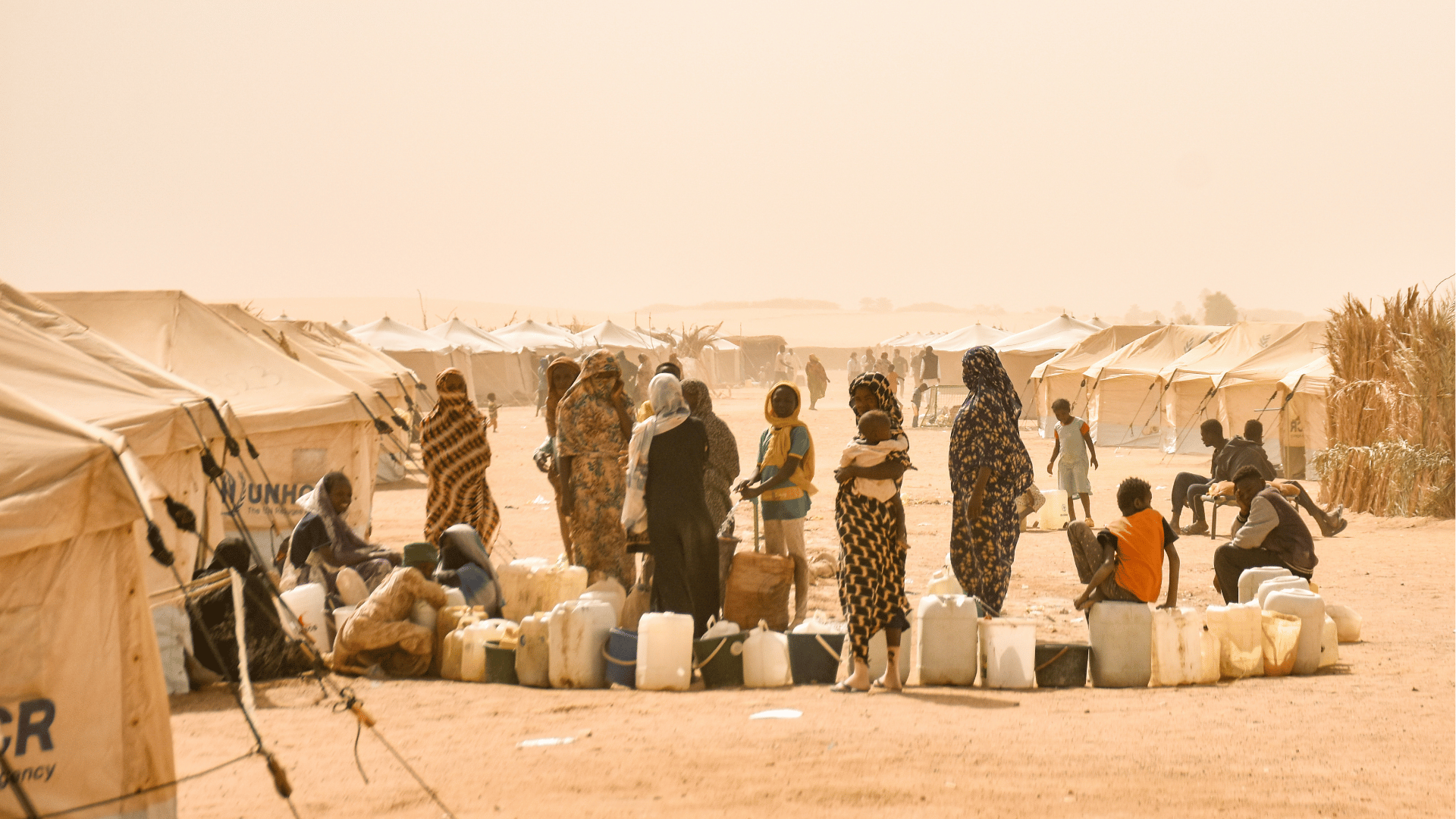 Pessoas, entre adultos e crianças, em um campo de refugiados no Sudão, reunidas entre tendas e recipientes de água em meio a um cenário árido e poeirento.