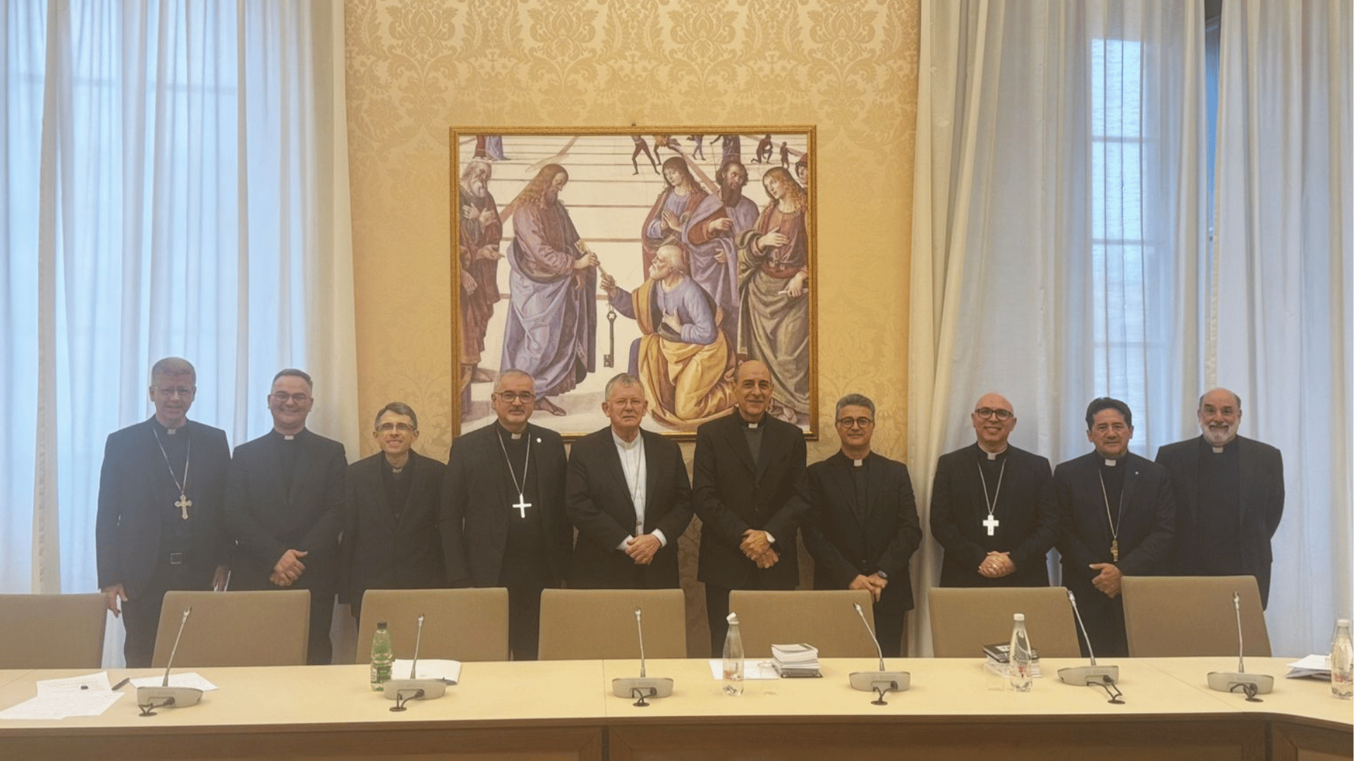 A imagem ilustra uma fila de dez religiosos, entre bispos e padres, atrás de uma mesa com cadeiras dispostas.
