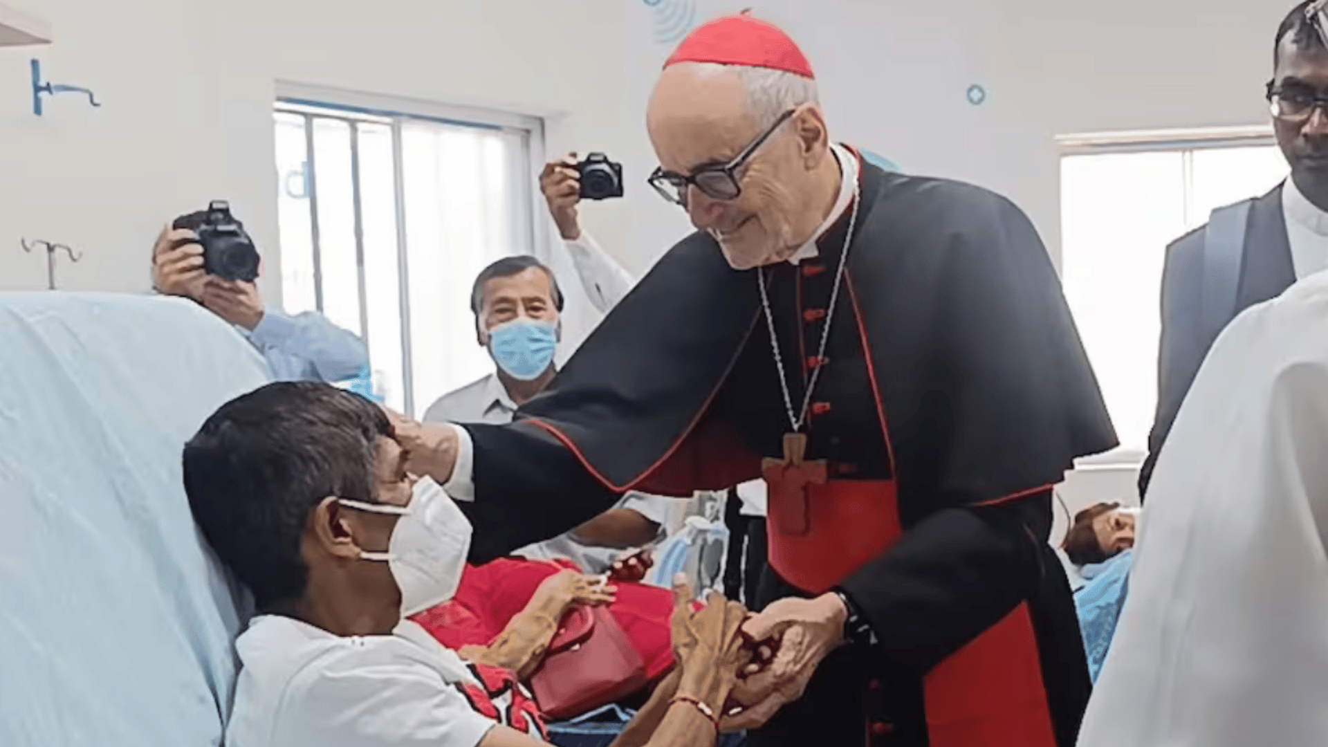 A imagem ilustra um cardeal da Igreja Católica, com suas habituais vestes, cumprimentando um homem enfermo, usando máscara, deitado em um leito hospitalar.