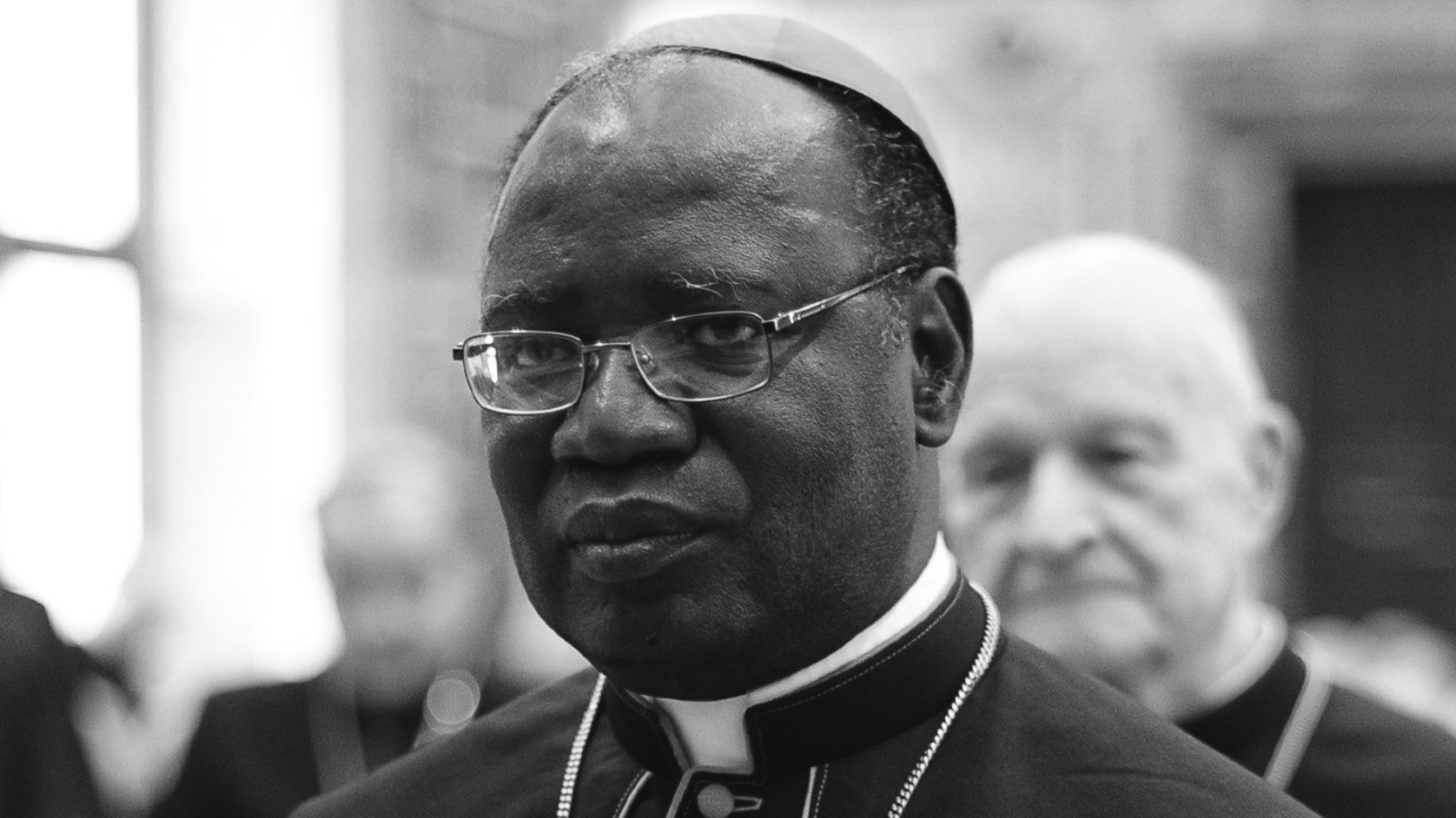 A imagem, em preto e brango, ilustra um cardeal da Igreja Católica, de pele negra e usando óculos.