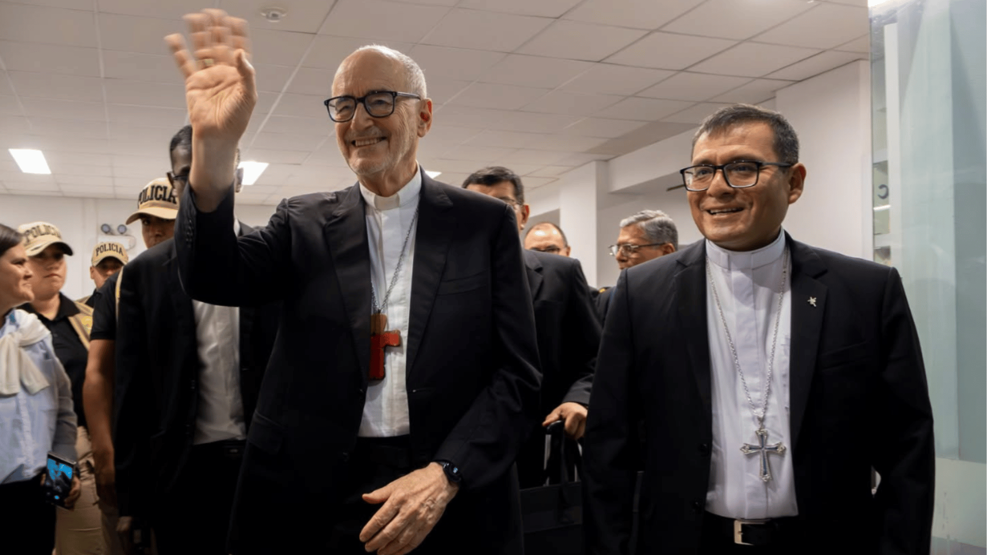 A imagem ilustra um cardeal da Igreja Católica vestindo clérgima e paletó com a mão direita erguida em gesto de aceno. Ao seu lado está um bispo católico, também vestindo clérgima e paletó.