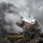 A imagem mostra o que parece ser um prédio ou casa destruído, com muita fumaça e destroços ao redor
