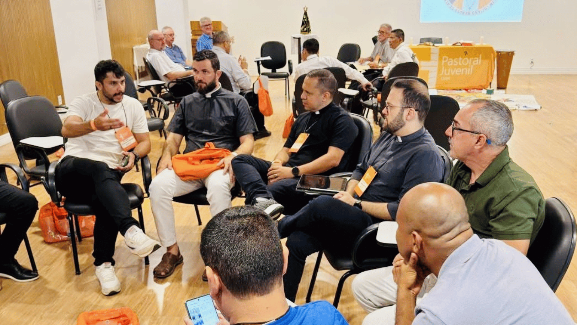 A imagem ilustra um grupo de homens, entre leigos e padres, sentados em círculo e conversando durante uma reunião. Ao fundo vê-se um outro grupo, disposto da mesma maneira.