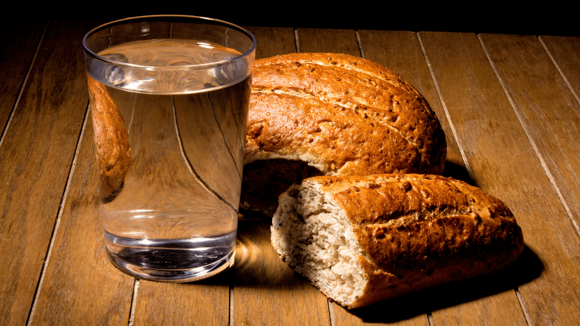 Jejum, abstinência e penitência A imagem ilustra um copo d'água e alguns pães sobre uma mesa de madeira, em referência ao jejum de pão e água.