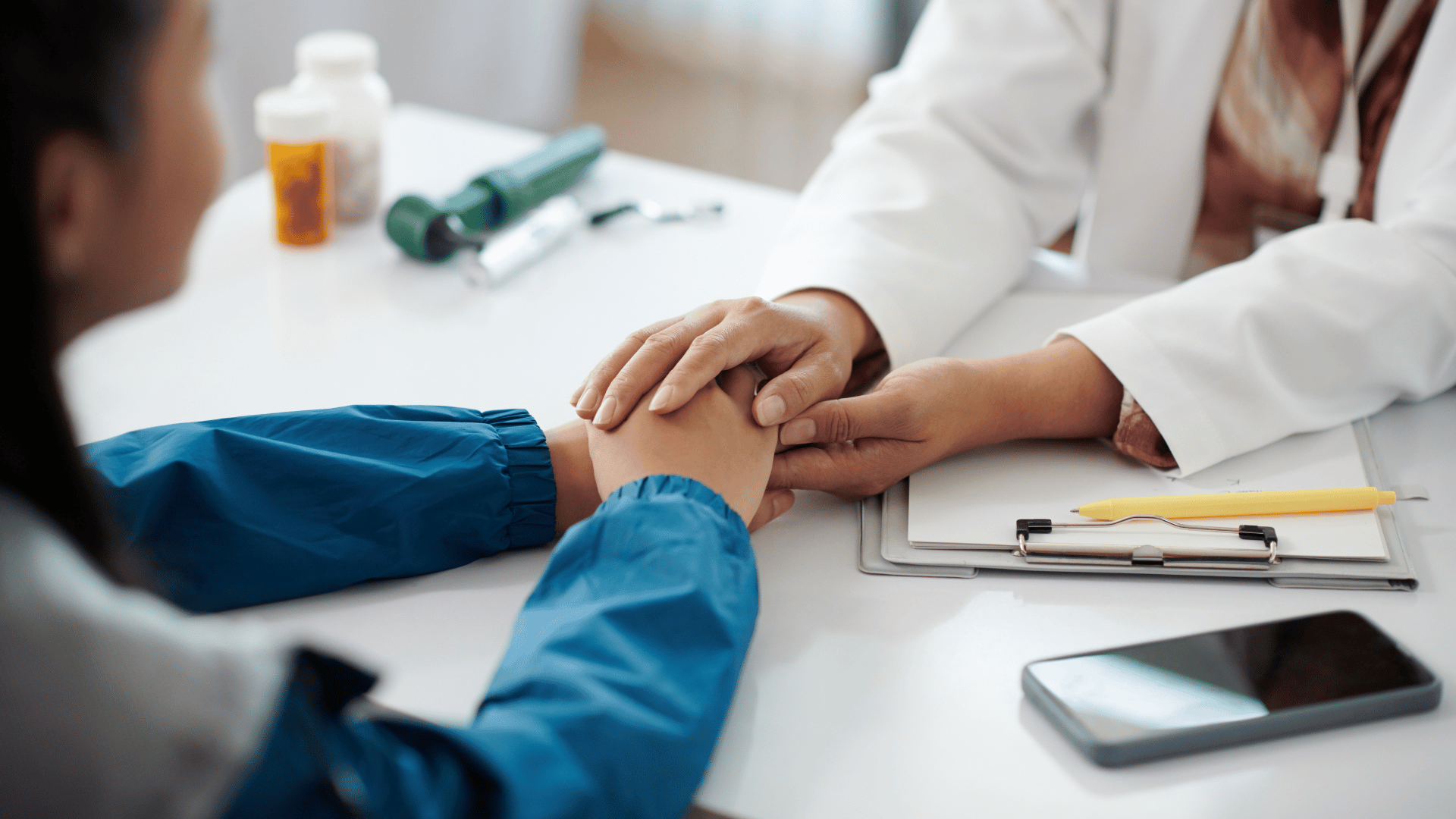 A imagem ilustra uma paciente e uma médica com as mãos sobrespostas sobre uma mesa com outros elementos médicos, em um gesto de acolhimento.