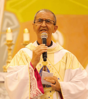 A imagem ilustra um padre católico, de pele parda e cabelos curtos e grisalhos, vestindo paramentos litúrgicos brancos e usando óuclos, com um microfone na mçao direita e um papel na mão esquerda.