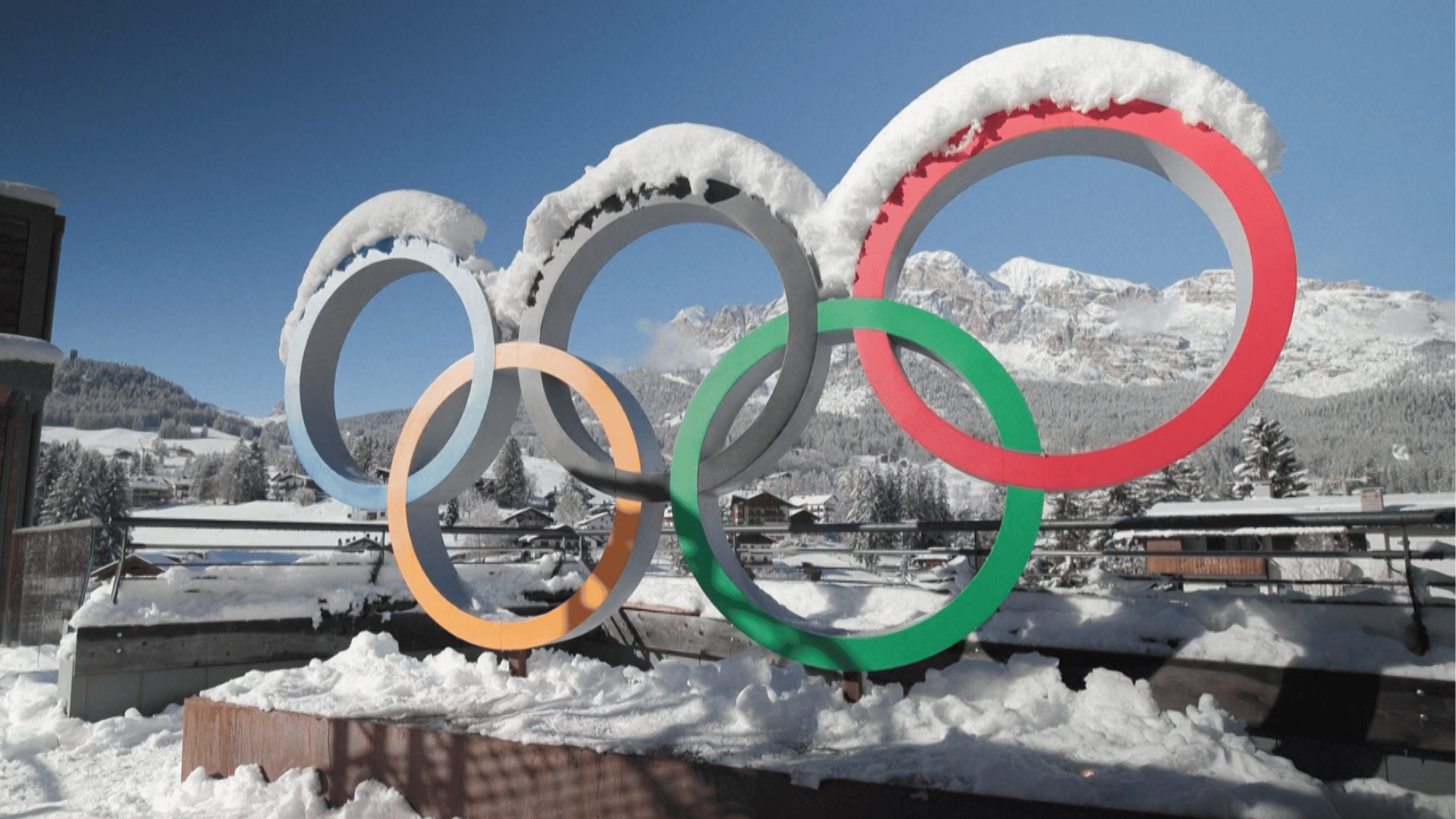 Olimpíadas de Inverno A imagem ilustra um display dos aros olímpicos, símbolo das Olimpíadas, cobertos por neve com um cenário montanhoso ao fundo.