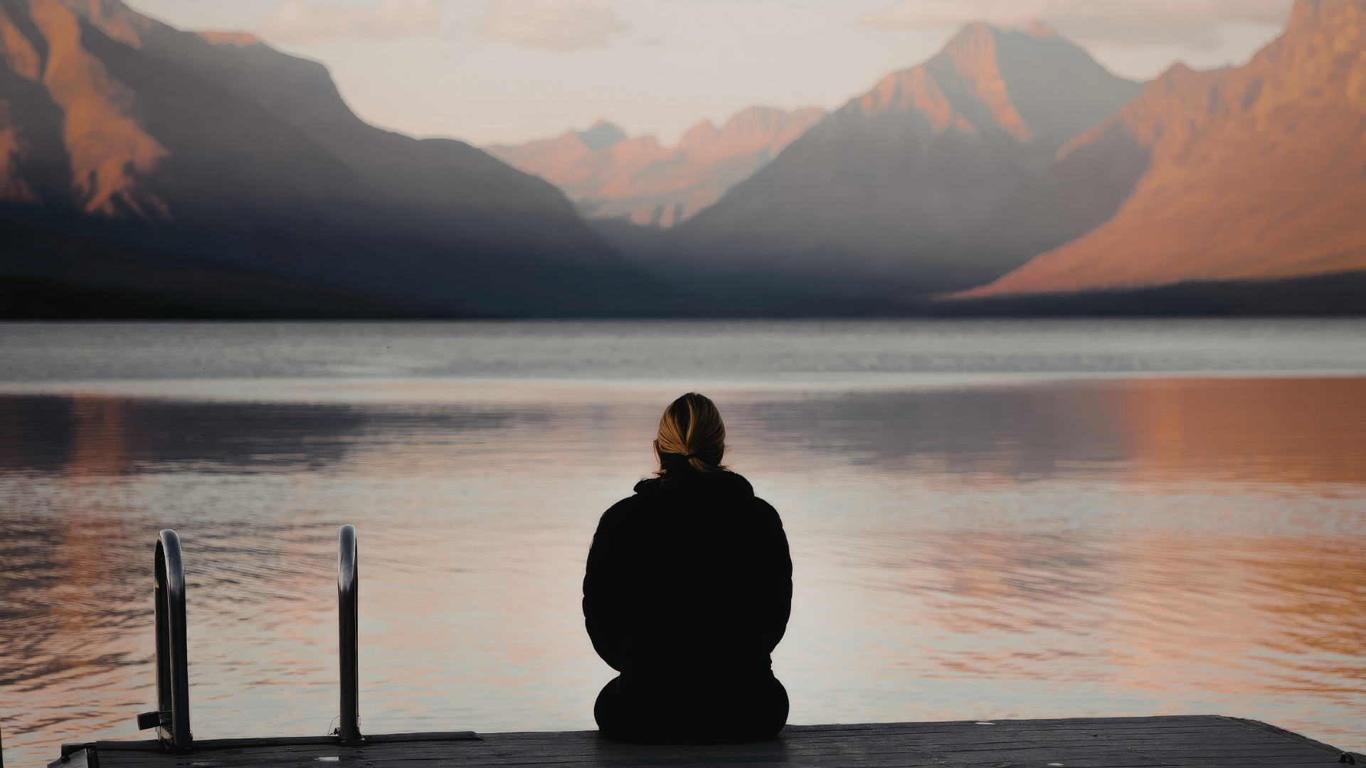 A imagem ilustra uma mulher vestindo agasalho preto, de costas, sentada em um deque à beira de um lago enquanto olha as montanhas ao fundo.
