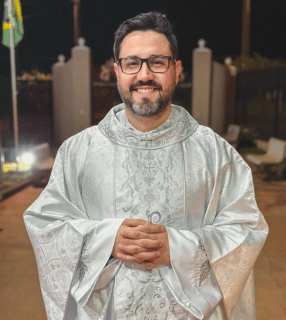 A imagem ilustra um padre católica, de pele branca, cabelo preto e barba e bigode grisalhos, vestido com uma casula branca.