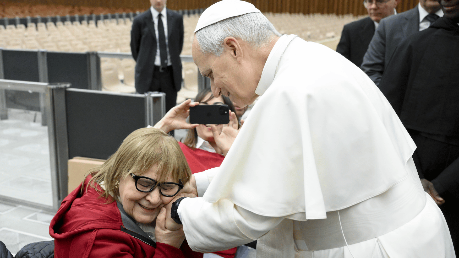 Uma senhora, sentada, sorri enquanto abraça, próximo ao seu rosto, as mãos do Papa Leão XIV, que aparece em pé
