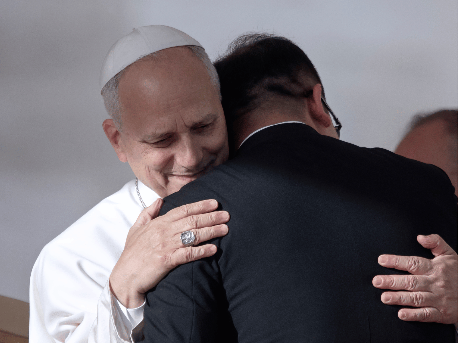 A imagem ilustra o Papa Leão XIV, com suas habituais vestes pontifícias, abraçando um padre católico.