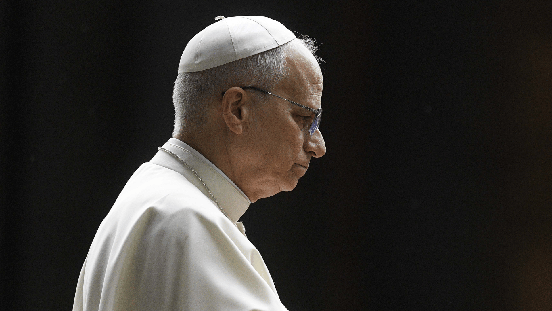 A imagem ilustra o Papa Leão XIV de perfil, com suas habituais vestes pontifícias e uma expressão séria, contrastando com o fundo escuro.