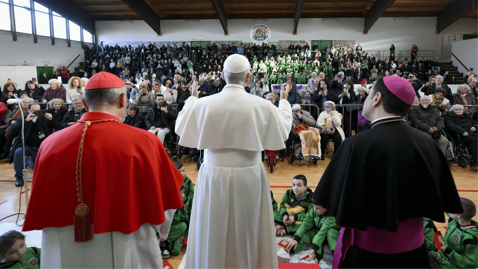 A imagem ilustra o Papa Leão XIV, ao centro, acompanhado por um cardeal à sua esquerda e um monsenhor à sua direita. Eles estão de costas, voltados para um grupo de pessoas sentadas na arquibancada de um ginásio.