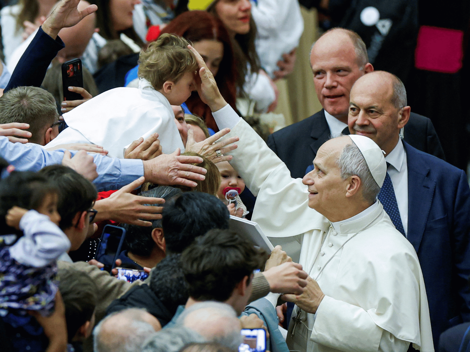 A imagem ilustra o Papa Leão XIV, com usas habituais vestes pontifícias, com o braço direito erguido para tocar a cabeça de uma criança e abençoá-la em meio aos fiéis.