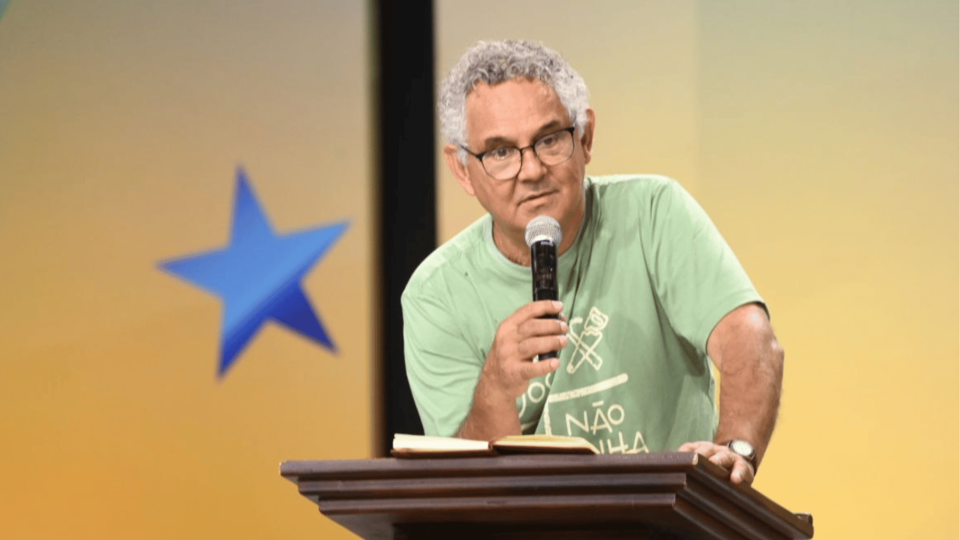 Diácono Nelsinho Corrêa no Rebanhão Canção Nova A imagem ilustra um homem de pele clara e cabelos cacheados grisalhos, vestindo uma camisa verde, como um microfone na mão direita.