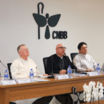 a presidência da CNBB aparece sentada à uma mesa atrás da qual, na parede, aparece o logo institucional da conferência