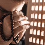 Homem em atitude de oração segura um terço entre as mãos, com a cabeça inclinada, em ambiente iluminado por luz suave.