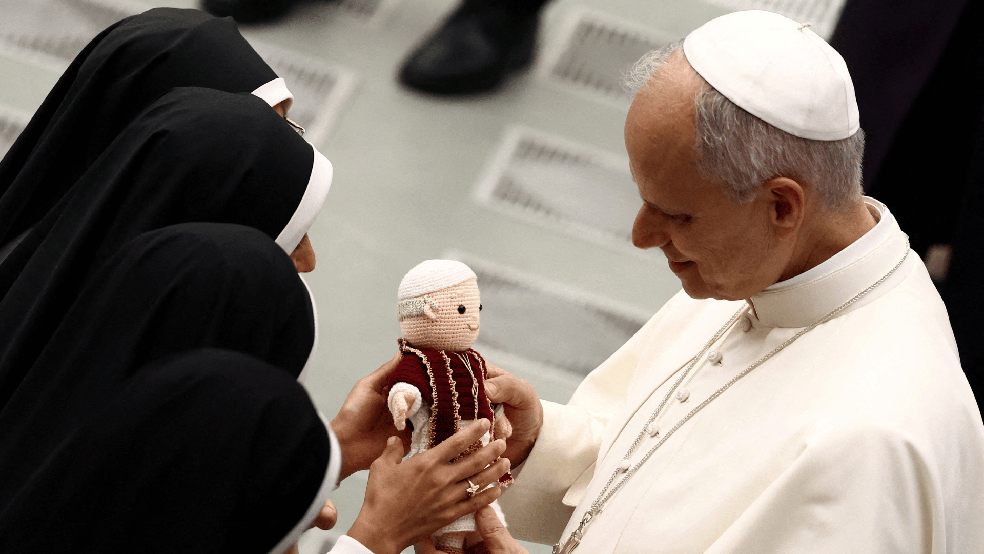 A imagem ilustra o Papa Leão XIV, com suas habituais vestes pontifícias, recebendo um boneco que o representa de um grupo de freiras.