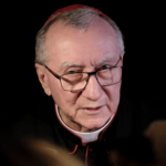 Cardeal Pietro Parolin, usando solidéu vermelho e vestes clericais pretas com detalhes vermelhos, falando durante entrevista, com fundo escuro.