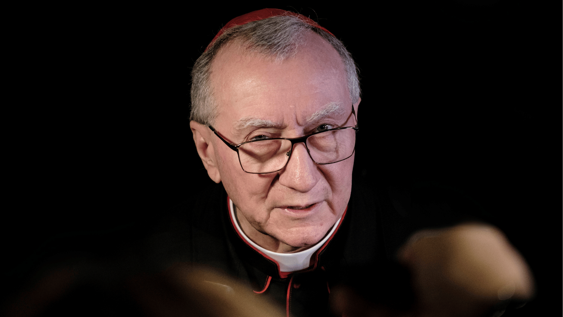 Cardeal Pietro Parolin, usando solidéu vermelho e vestes clericais pretas com detalhes vermelhos, falando durante entrevista, com fundo escuro.