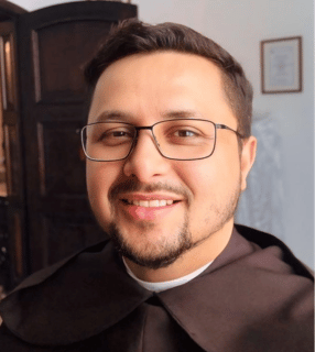 A imagem ilustra um homem branco de cabelos castanhos e barba e bigode curtos, usando óculos e hábito religioso.