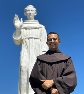 A imagem ilustra um frei franciscano, com o tradicional hábito marrom, e uma imagem branca de Frei Galvão ao fundo.