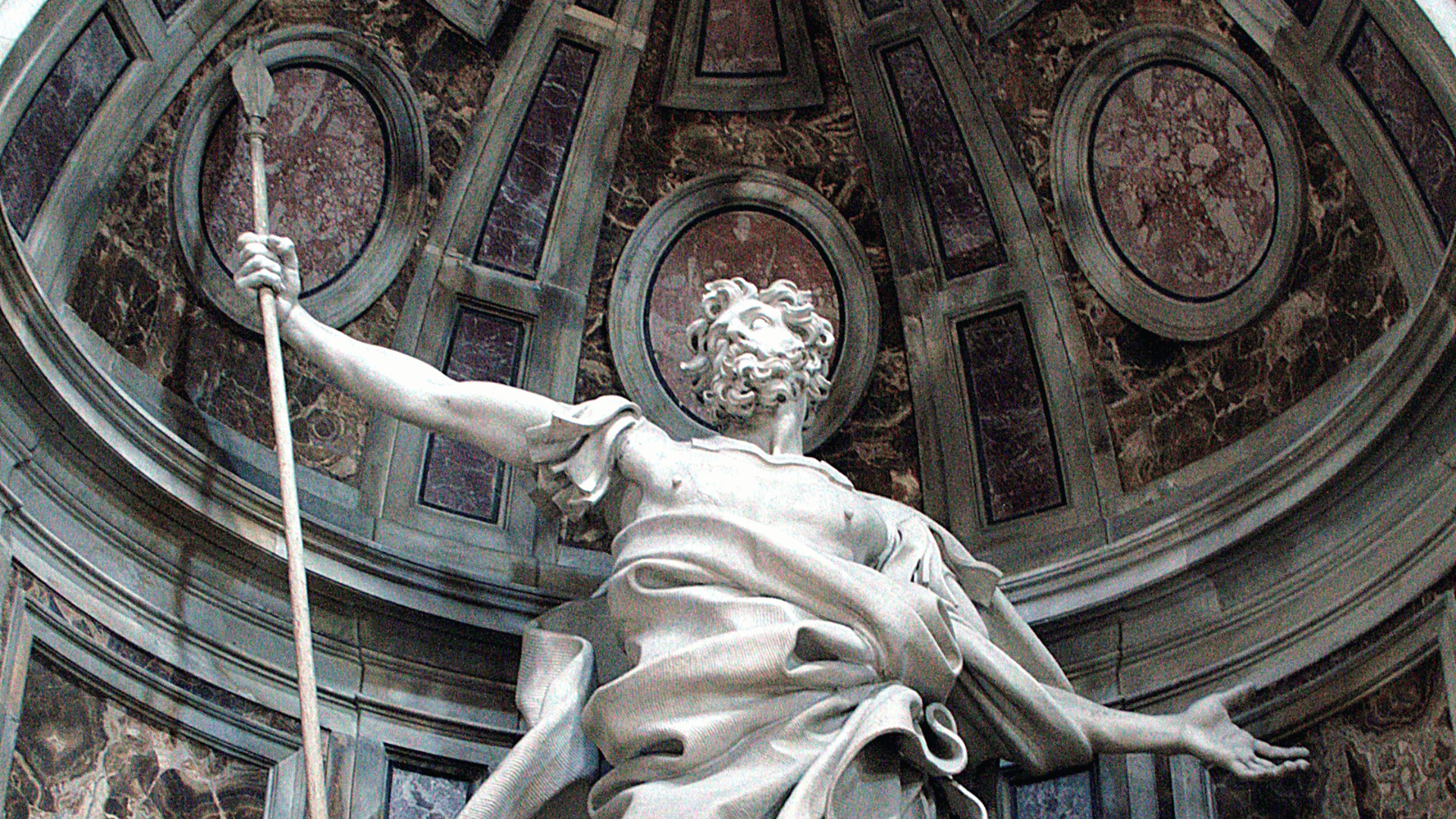 A foto ilustra uma estátua de São Longuinho, que porta uma lança na mão direita e tem o braço esquerdo estendido para o lado. Ao fundo, vê-se uma cúpula.