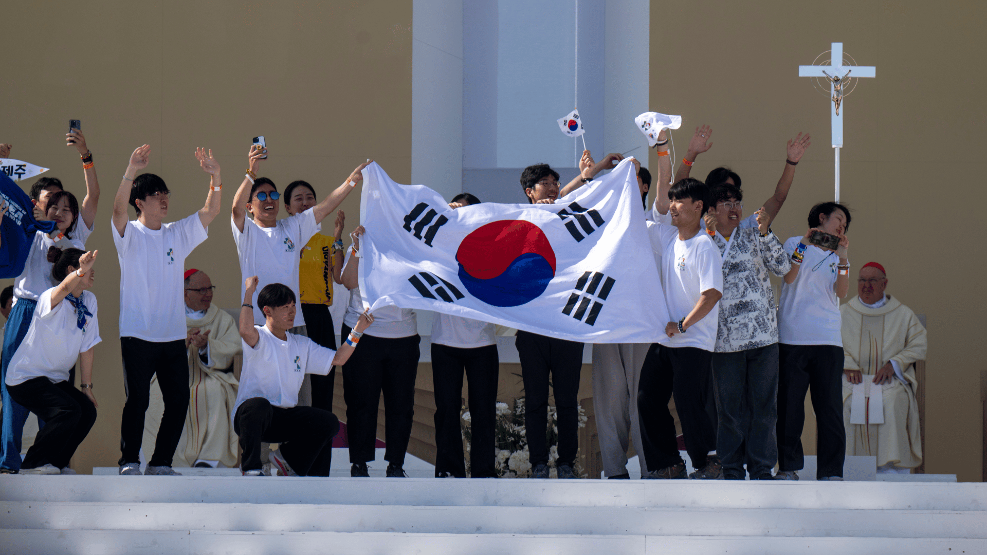 A imagem ilustra um grupo de jovens segurando e agitando uma bandeira grande e algumas bandeiras menores da Coreia do Sul.
