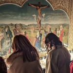 Fiéis rezam diante de imagem de Jesus crucificado, com velas acesas, em ambiente de oração dentro de igreja.