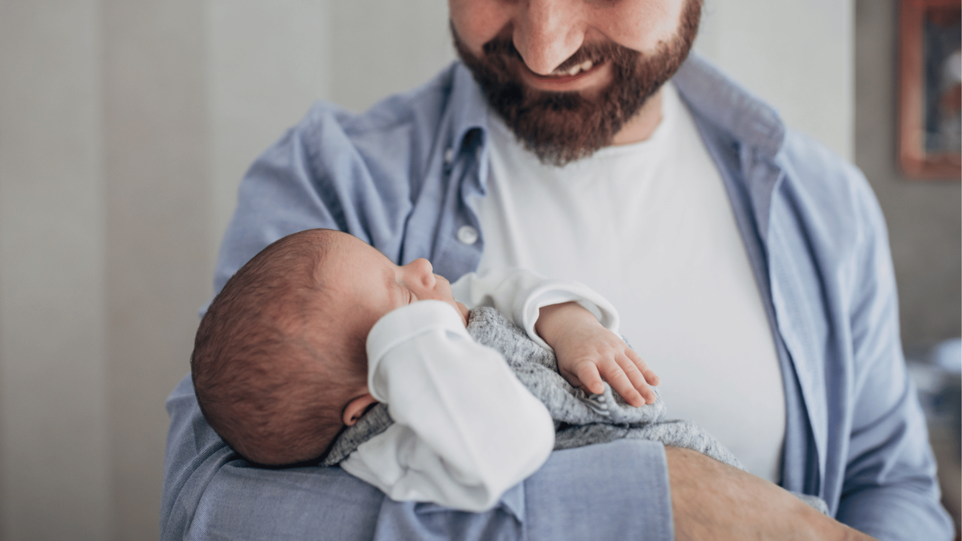 Pai segurando um bebê recém-nascido nos braços, olhando para a criança com carinho em ambiente interno.