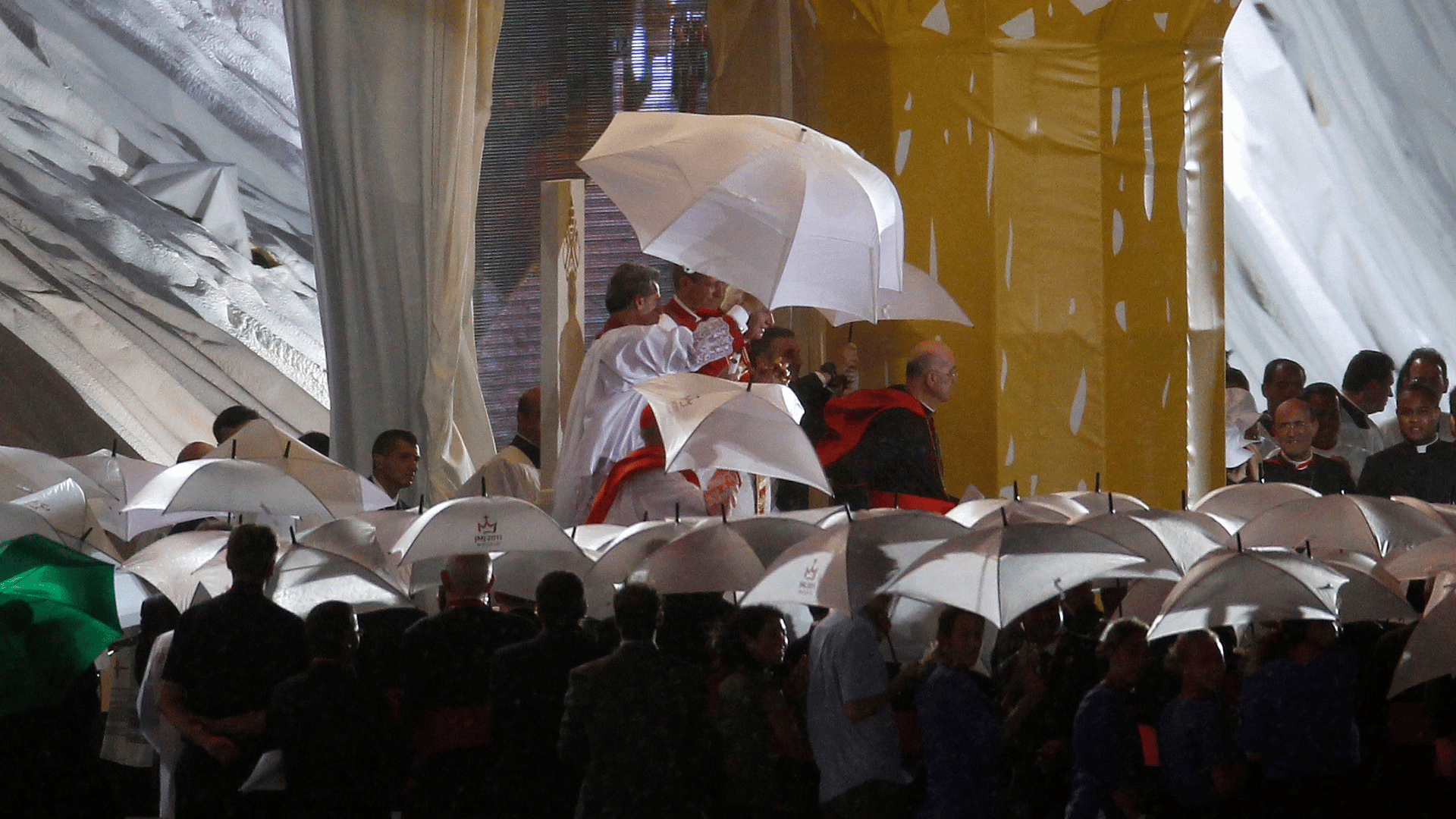 A imagem ilustra o Papa Bento XVI em meio a guarda-chuvas para se proteger de tempestade na JMJ Madri 2011. É possível ver ainda outras pessoas com guarda-chuvas na imagem.