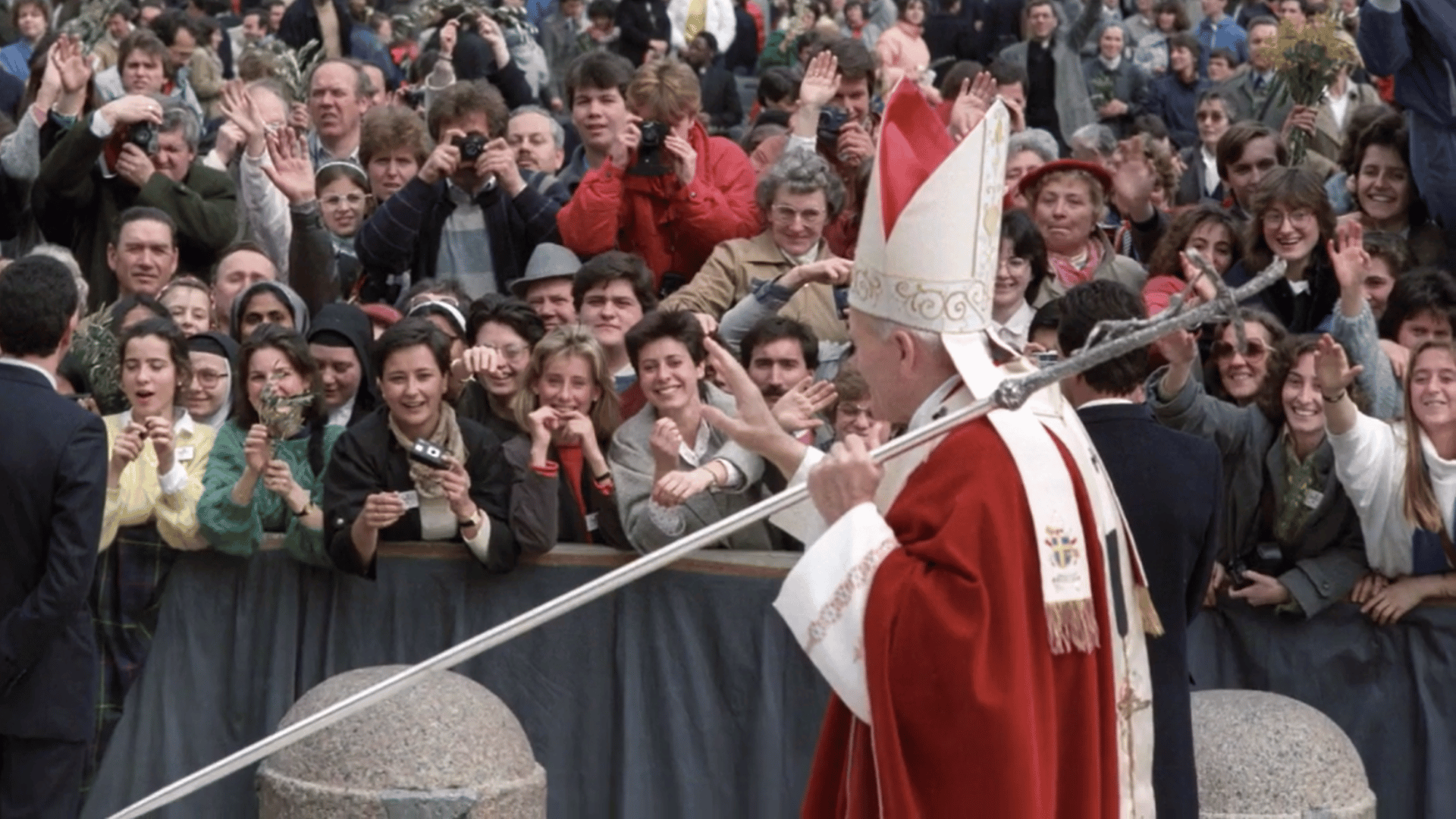 A imagem ilustra o Papa João Paulo II, com paramentos litúrgicos vermelhos e a férula papal na mão esquerda, acenando para os peregrinos reunidos na Praça São Pedro.
