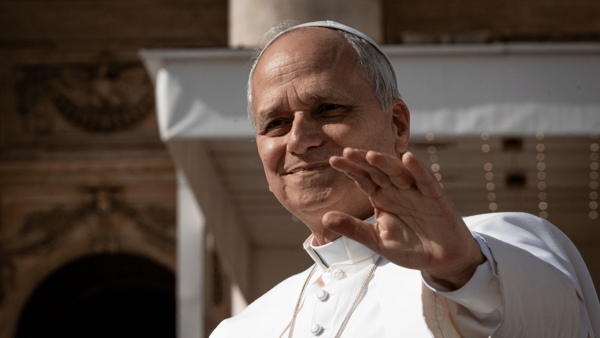 Papa Leão XIV A imagem ilustra o Papa Leão XIV, com suas habituais vestes pontifícias, uma expressão neutra e a mão esquerda estendida para a frente em gesto de saudação.